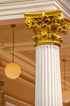 An ornate Corinthian column with a decorative gold capital stands beneath a coffered ceiling with pendant lights.