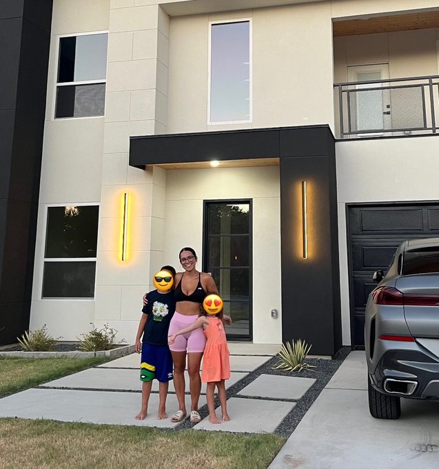 A family photo taken outside a modern two-story home with vertical light fixtures flanking the front door; a woman and two children pose together on a concrete walkway, with their faces covered by emoji stickers.