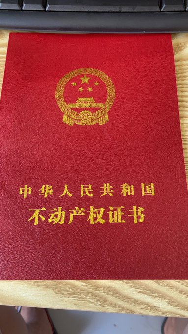 A red Chinese property ownership certificate booklet featuring the national emblem and gold text reading '中华人民共和国 不动产权证书' (People's Republic of China Real Estate Ownership Certificate).
