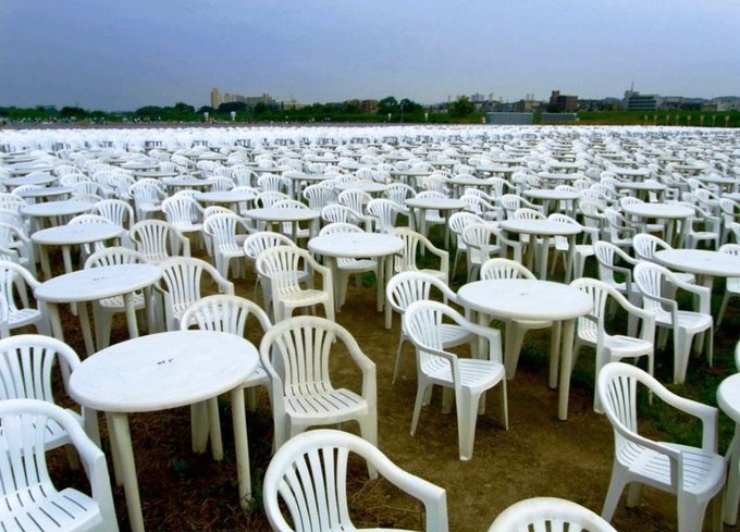 A vast field filled with hundreds of white plastic tables and chairs arranged in neat rows under a cloudy sky, with buildings visible in the distance.