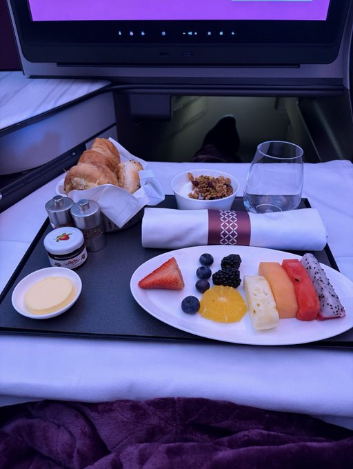 An airline meal service on a tray featuring a fruit plate with strawberries, blueberries, blackberries, pineapple, cantaloupe, and dragon fruit, alongside bread rolls, a granola bowl, a water glass, and condiments, set in front of a seatback screen.