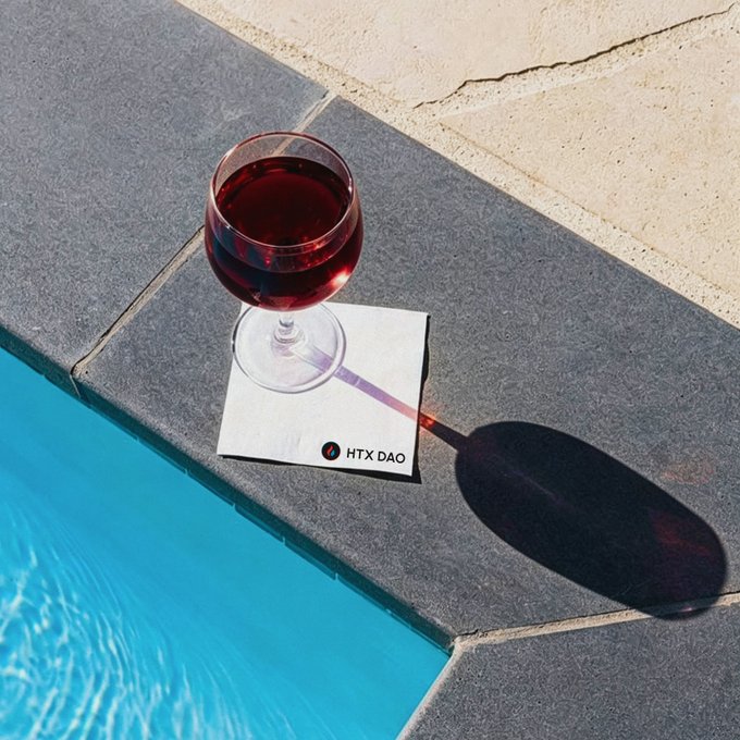 A glass of red wine rests on a napkin branded "HTX DAO" beside a blue swimming pool, with sunlight casting a long shadow across the stone tiles to evoke the post's "GM" theme of sunny, leisurely moments by the water.