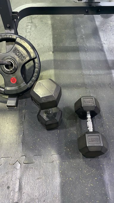 A gym floor with black rubber mats showing two black hex dumbbells labeled '15KG' lying on the ground near a weight plate rack holding a '20KG' plate.