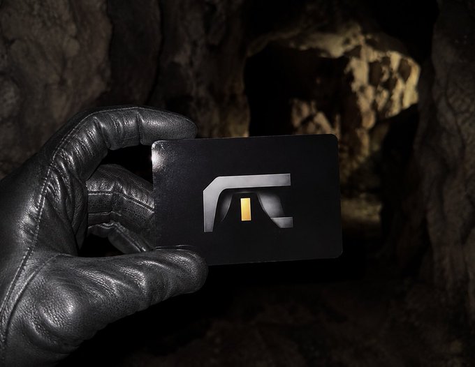A gloved hand holds up a black card featuring a silver logo with a small gold rectangle, set against a dark, cave-like background with rocky textures.