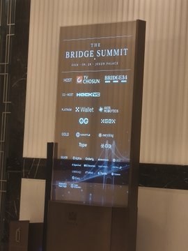 An illuminated digital display showing 'THE BRIDGE SUMMIT 2024 · IN JA · TOKYO PALACE' along with sponsor and partner logos including WEMIX, CHOSUN, HORIZON3, WALLET, OK, XBN, SUI, and others.