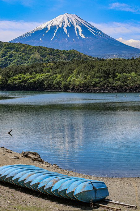 A scenic view of Mount Fuji snow-capped peak rising behind a forested hillside and a calm blue lake, with a blue upturned boat resting on the shore in the foreground.