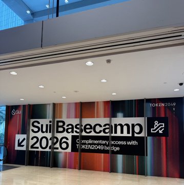 A modern lobby space features a large colorful banner advertising "Sui Basecamp 2026" with the tagline "Complimentary access with TOKEN2049 badge," alongside the Sui logo and TOKEN2049 branding. The display ties directly to the post's theme by promoting the upcoming Sui Basecamp event as a hub for sharing, connecting, and creating value within the Sui ecosystem.