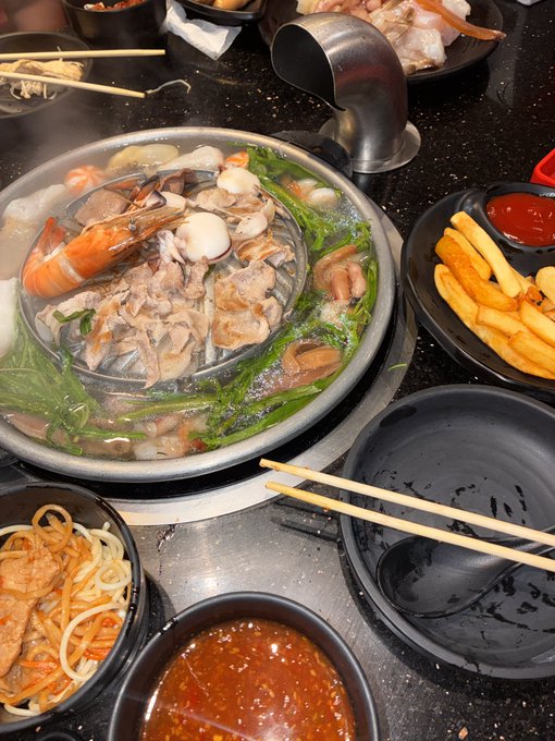 A top-down photo of a hot pot meal featuring meat, shrimp, clams, and greens cooking in a divided pot, surrounded by side dishes like french fries, noodles, and dipping sauces.