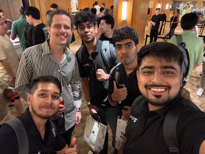 A group selfie of five smiling men at an indoor event, with several wearing black 'INDEED' branded shirts and one holding a beverage.