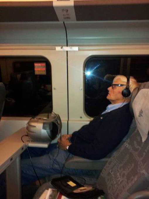 urn of water sits on a table, accompanied by a man wearing headphones and a blue jacket, with a window offering a view of the train's interior.