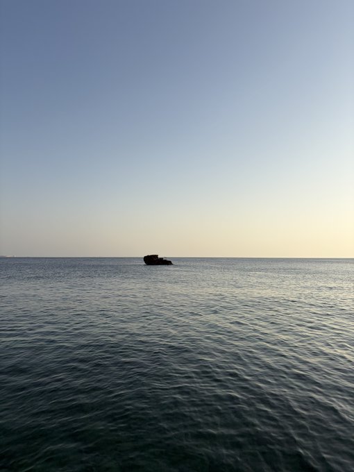 Calm seascape photograph showing a small rock formation in the middle of the ocean under a clear gradient sky transitioning from blue to soft yellow near the horizon.