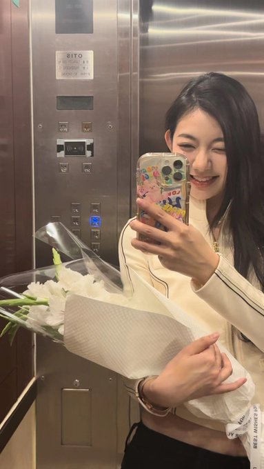 A smiling young woman with long dark hair takes a mirror selfie in an elevator while holding a bouquet of white flowers wrapped in paper.