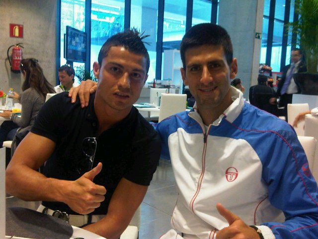 A photograph of two men at an indoor event, both giving a thumbs-up, with one wearing a black shirt and the other in a blue and white jacket with a logo.