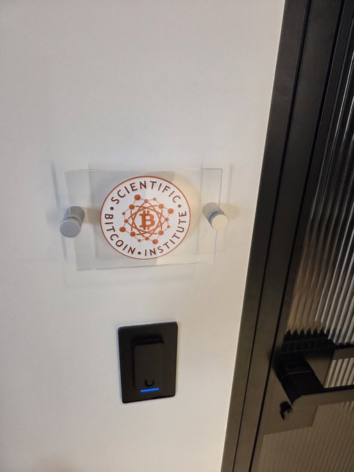 urn of gold and red, with a white circle and "Scientific Bitcoin Institute" logo on it, is mounted on a black door frame.