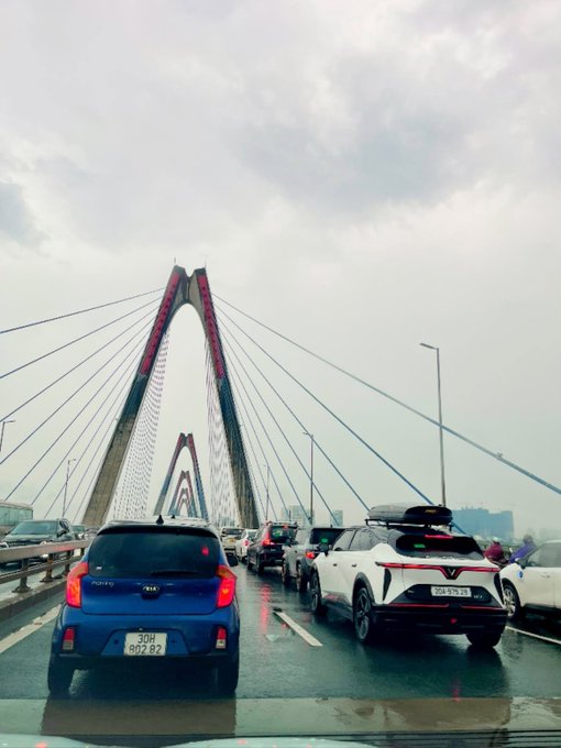 A view from a car on a rainy day shows traffic on a large cable-stayed bridge, with a blue Kia '30H 902.82' in front and a white SUV with a roof box nearby.