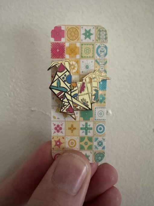 A hand holds a colorful patterned card or ticket with an origami crane pin attached to it.