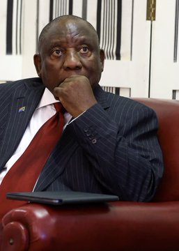A photograph of South African President Cyril Ramaphosa seated on a red leather chair, wearing a dark pinstripe suit and red tie, with his hand resting thoughtfully against his chin.