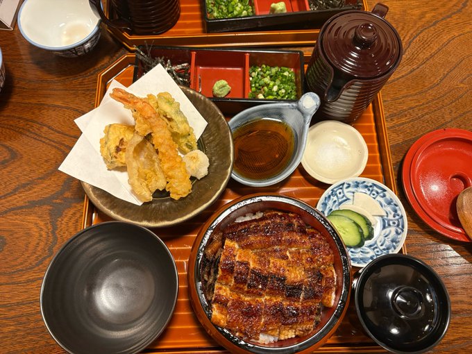 An overhead shot of a Japanese meal set featuring a bowl of unagi (grilled eel) over rice, tempura, dipping sauce, wasabi, pickles, and various small dishes on a wooden tray.