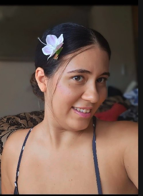 A smiling young woman with dark hair pulled back, adorned with a light purple orchid flower hair clip, wearing a dark top with thin straps.