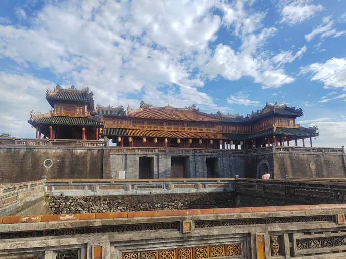 A traditional East Asian palace or temple complex with ornate tiled roofs and red columns, viewed from behind an intricately carved stone balustrade under a bright blue sky with scattered clouds.