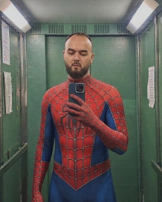 A bearded man taking a mirror selfie in an elevator while wearing a red and blue Spider-Man costume, holding a smartphone to capture the photo.