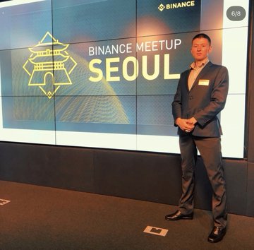 The image shows a man in a suit standing confidently in front of a "BINANCE MEETUP SEOUL" backdrop, illustrating his humble beginnings in South Korea's crypto scene around 2018 before his rise to a $1.5M overnight success and Dubai lifestyle. The Binance event photo represents an early step in his 9-year crypto journey from obscurity to influencer status with 1M+ followers.