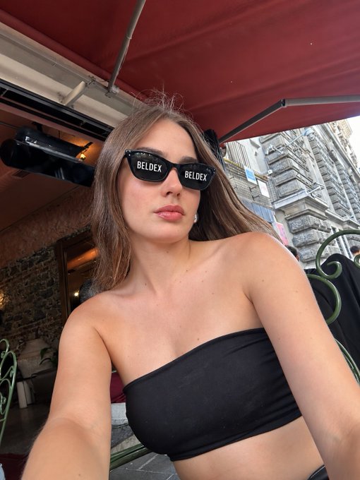 A selfie of a woman wearing black sunglasses with 'BELDEX' written across them and a black strapless top, sitting outdoors under a red awning.