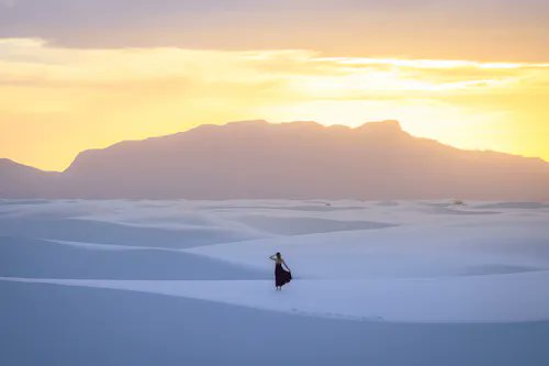 A solitary figure draped in flowing black stands atop vast, undulating white sand dunes, silhouetted against a dramatic mountain range bathed in the warm golden glow of a setting or rising sun. The image perfectly captures the "Lone Wanderer" theme, emphasizing the small human presence amid the immense, serene desert landscape.