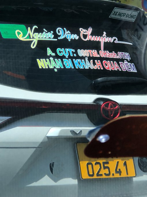 The rear of a white Toyota vehicle displays colorful Vietnamese text on the window reading 'Người Vận Chuyển' and 'NHẬN ĐI KHÁCH QUA BẾN', with a yellow license plate showing '025.41'.