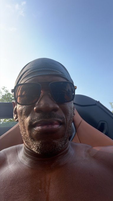 A man wearing a gray durag and sunglasses takes a relaxed selfie in a convertible against a clear blue sky, his sun-drenched, no-shirt look embodying a laid-back, summer-cruising vibe. The carefree, "ready for the warm season" attitude mirrors the optimism of "How bad do you want the bull market?"—capturing a mood of sun-soaked confidence and bullish energy.