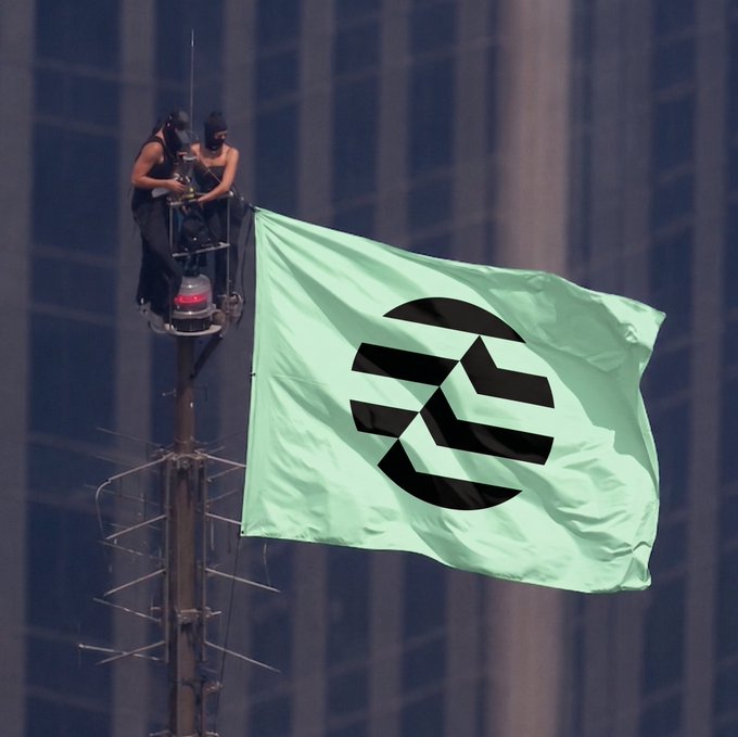 Two people are perched at the top of a pole, unfurling a large green flag featuring a black geometric logo resembling an Ethereum-style symbol against a cityscape backdrop. The caption "Worth the view" highlights the daring, high-altitude spectacle of raising the crypto-themed flag atop a towering structure.