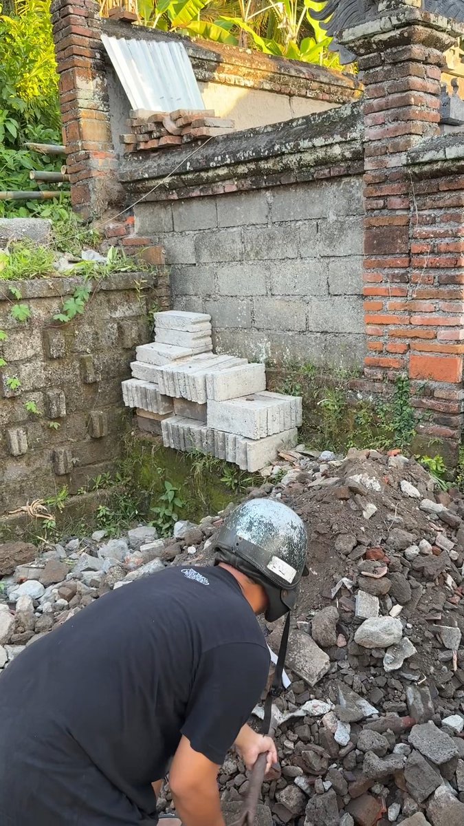 A construction worker wearing a hard hat is bent over working with rubble and debris in front of a partially demolished brick and concrete wall.