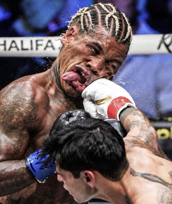 A close-up action shot of a boxer with braided hair taking a hit to the face during a match, with sweat and water spraying through the air.