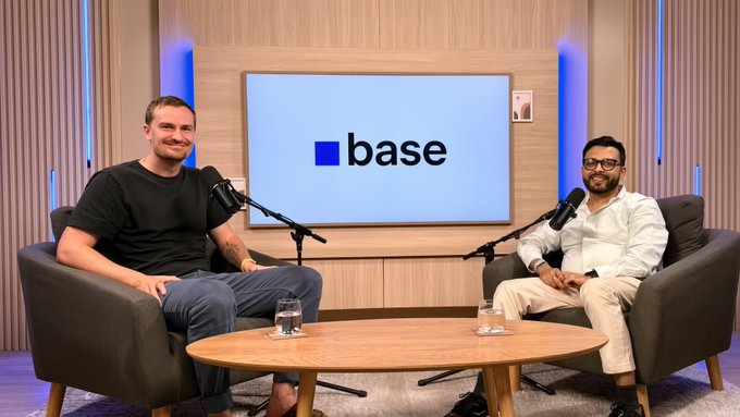 Two men seated in armchairs with microphones during a podcast interview, with a 'base' logo displayed on the monitor between them.