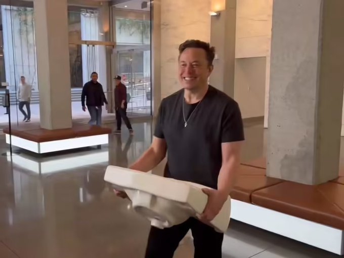 Elon Musk smiling while carrying what appears to be a rolled-up poster or item in a modern building lobby.