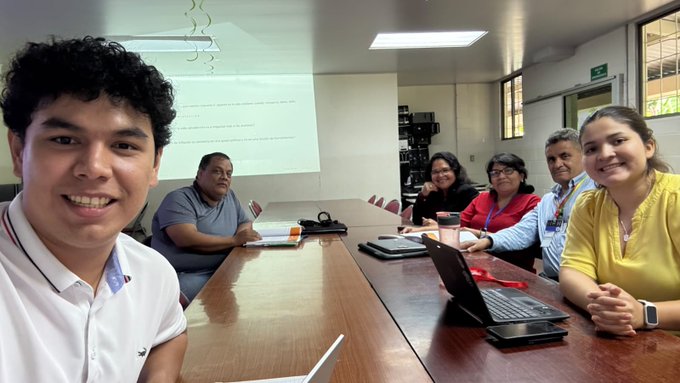 A group of educators smile together around a conference table during a Bitcoin Diploma 2.0 teacher training session, with a projector screen displaying presentation content behind them. The image perfectly illustrates the post's message about training teachers who will then educate entire classrooms about Bitcoin for years to come.
