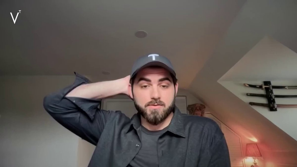 The image features a man wearing a black shirt, hat, and baseball cap, who appears to be posing for a photo or video call while holding his head up with one hand. The background of the image shows a white wall, which contrasts nicely with the man's dark attire. There is also an interesting detail in the image: a sword mounted on the wall behind the man.