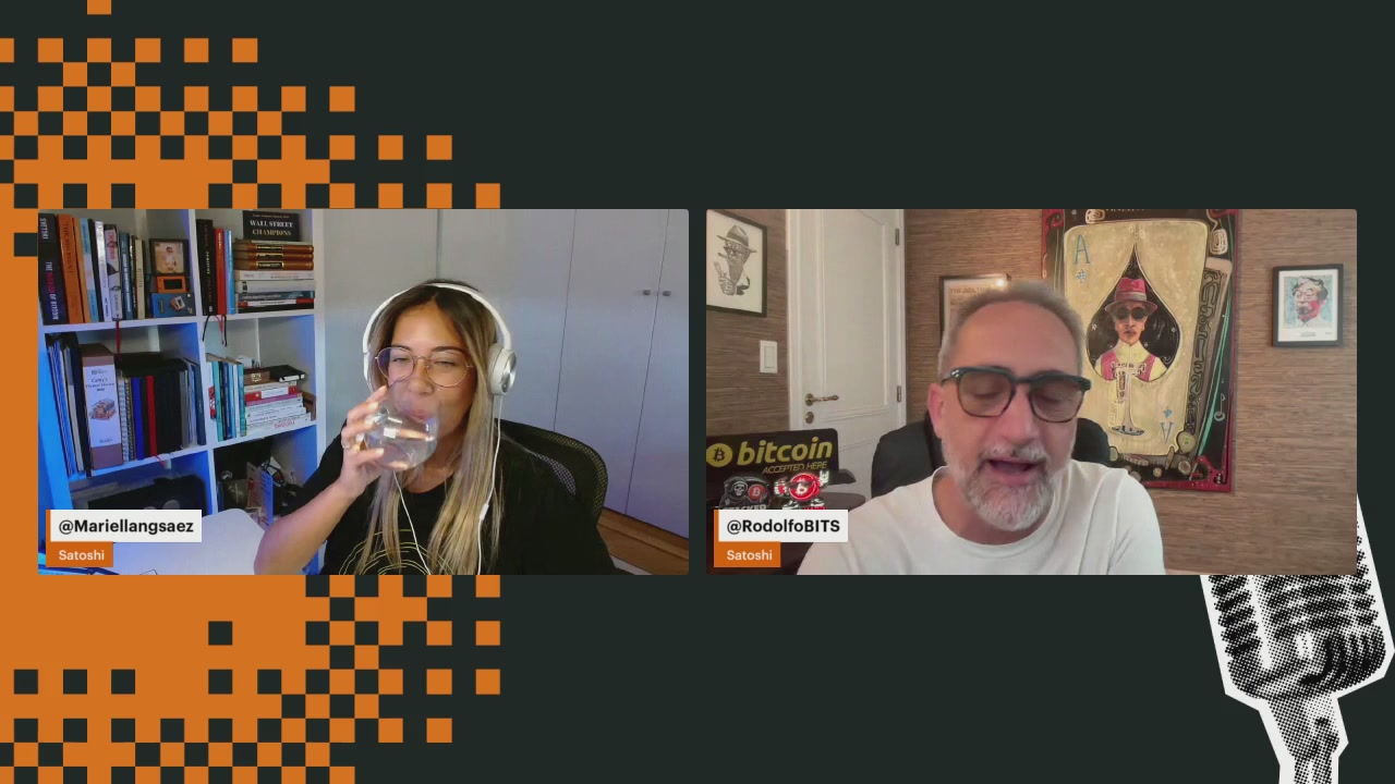 The image shows a man wearing glasses sitting behind a desk, with another person wearing headphones and drinking water next to him. The background features an orange and black checkered pattern. The caption of the image reads "Todos Somos Satoshi #8".