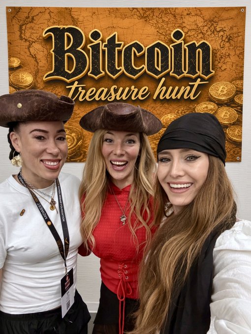 Three women in pirate hats smile in front of a 'Bitcoin treasure hunt' banner decorated with gold coins.