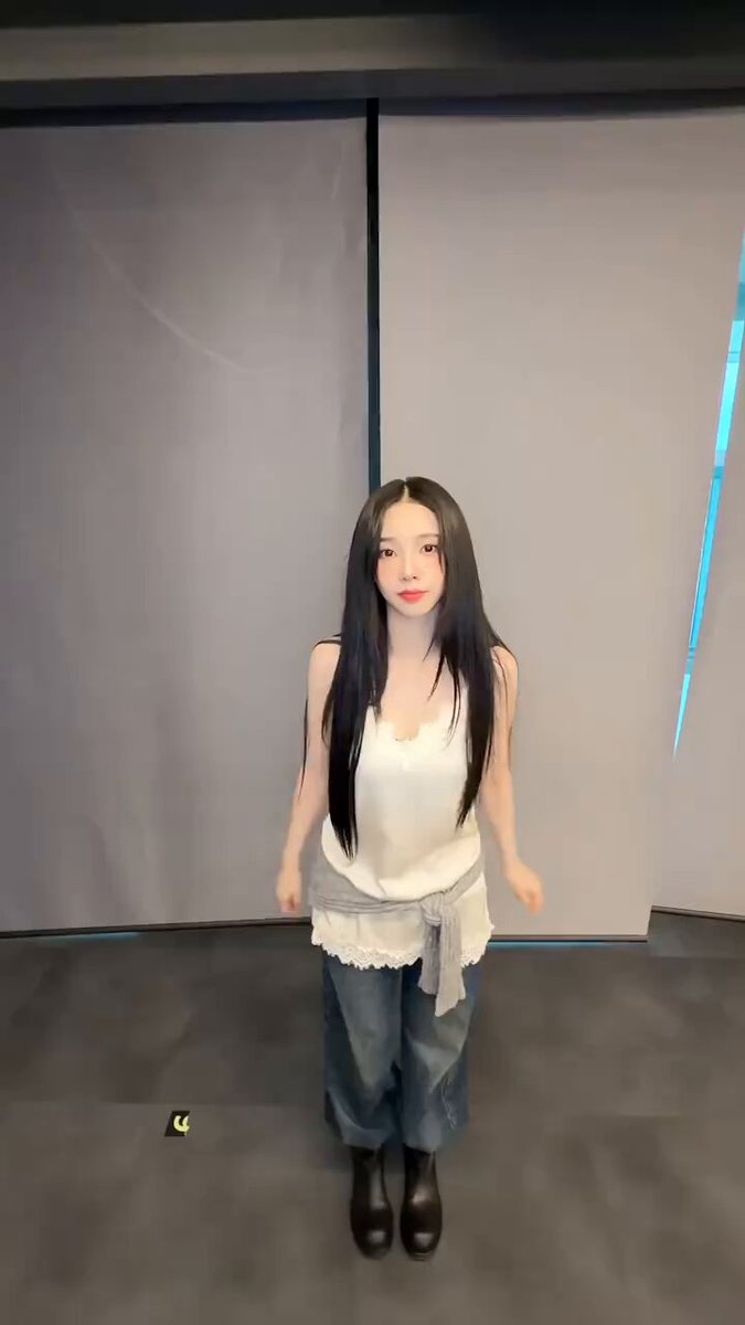 The image features a woman with long hair, dressed in blue jeans and a white top, standing in front of a gray wall. She appears to be posing for a photo or video, possibly for an online platform like Instagram. The background includes a window that is partially open, allowing some light into the room. There are also two small objects on the floor near her - a yellow object and a green one.