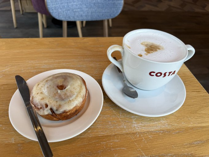 A Costa coffee cup with foam art sits on a wooden table next to a glazed cinnamon roll on a white plate with a knife.