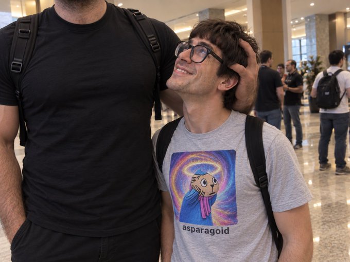 A shorter man wearing glasses and a gray shirt featuring an 'asparagold' graphic smiles as he leans against a taller person in a black shirt.
