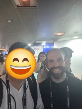 A selfie shows a smiling bearded man standing next to a person whose face is obscured by a large smiley emoji, with other people and a 'WELCOME' sign visible in the background.