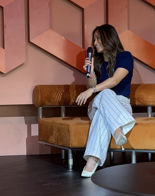 A woman with long brown hair, wearing a navy blue short-sleeve top and white pinstripe pants, sits on a rust-colored couch while holding a microphone, smiling as she speaks during the Cointelegraph CONNECT panel discussion on crypto liquidity, fragmentation, and AI.