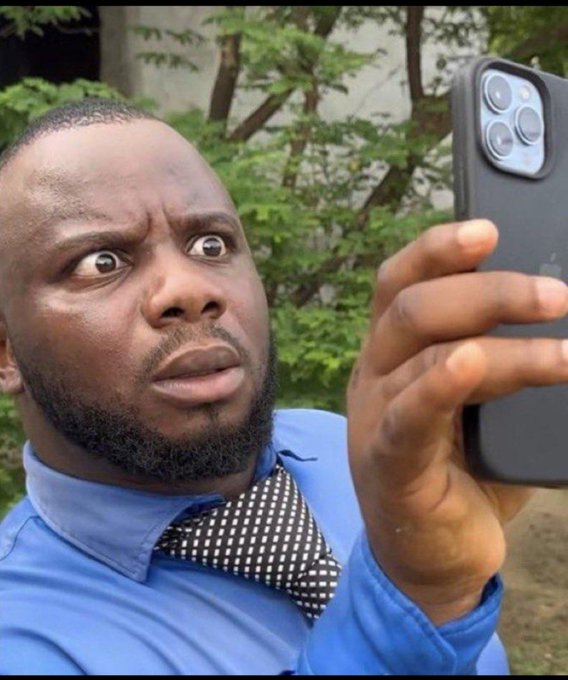 A man in a blue shirt and patterned tie holds up a smartphone, his face showing a shocked or wide-eyed surprised expression.