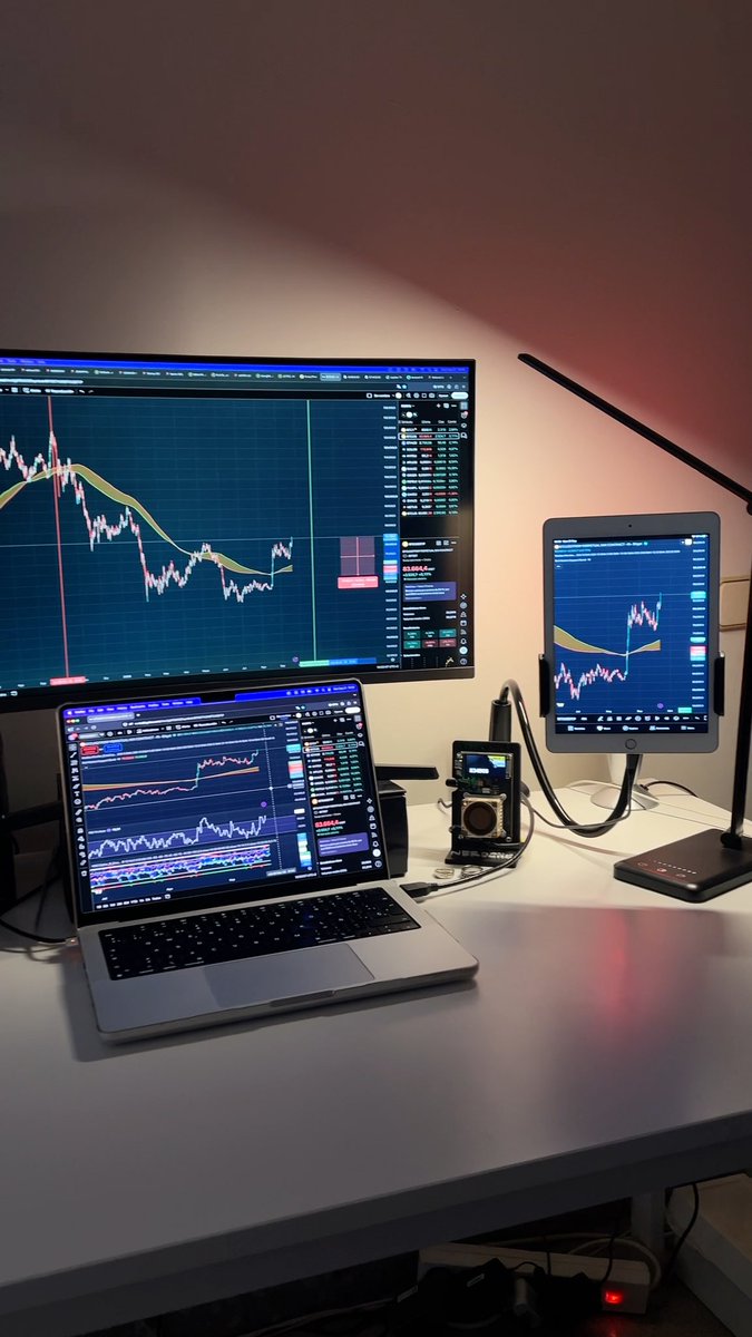 A dimly lit desk setup featuring a laptop, a large monitor, and a tablet, all displaying financial trading charts with candlesticks and indicators.