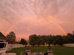 A scenic view of a complete rainbow arching over a suburban backyard with trees and a stone fire pit at sunset.