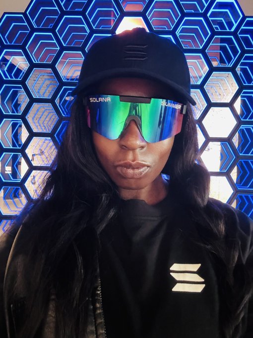 A confident woman poses in front of a futuristic blue hexagonal backdrop, wearing a black cap and mirrored sunglasses with "SOLANA" and "SUMMER" text on the temples, paired with a black jacket featuring a logo. The Solana branding ties directly into her message about building a "team of degenerates" willing to take calculated, asymmetric bets on unconventional ideas — likely within the Solana/Meteora crypto ecosystem she's promoting.