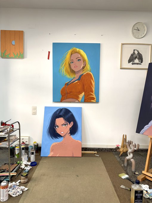 An artist's studio with two large anime-style paintings of female characters (one blonde, one with dark hair) leaning against a white wall, alongside paint supplies, a metal cart, a wall clock, and a framed bird print.