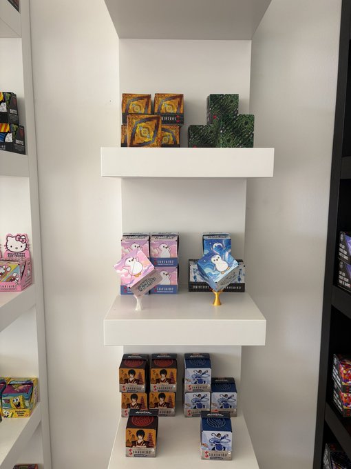 White retail display shelves showcase colorful Shashibo shape-shifting puzzle boxes in various designs including flowers, penguins, Hello Kitty, and astronaut themes, consistent with a visit to the Shashibo office on Venice Beach.